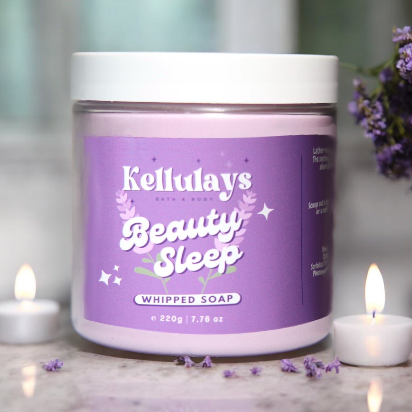 Image of a jar of Kellulays Bath & Body Beauty Sleep Whipped Soap. The jar is labeled with a purple sticker featuring the product name 'Beauty Sleep' in bold white font, accompanied by lavender and botanical illustrations. The jar contains 220g (7.76 oz) of whipped soap and has a white lid. The background includes soft-focus lavender sprigs and lit candles, creating a calming and serene atmosphere.