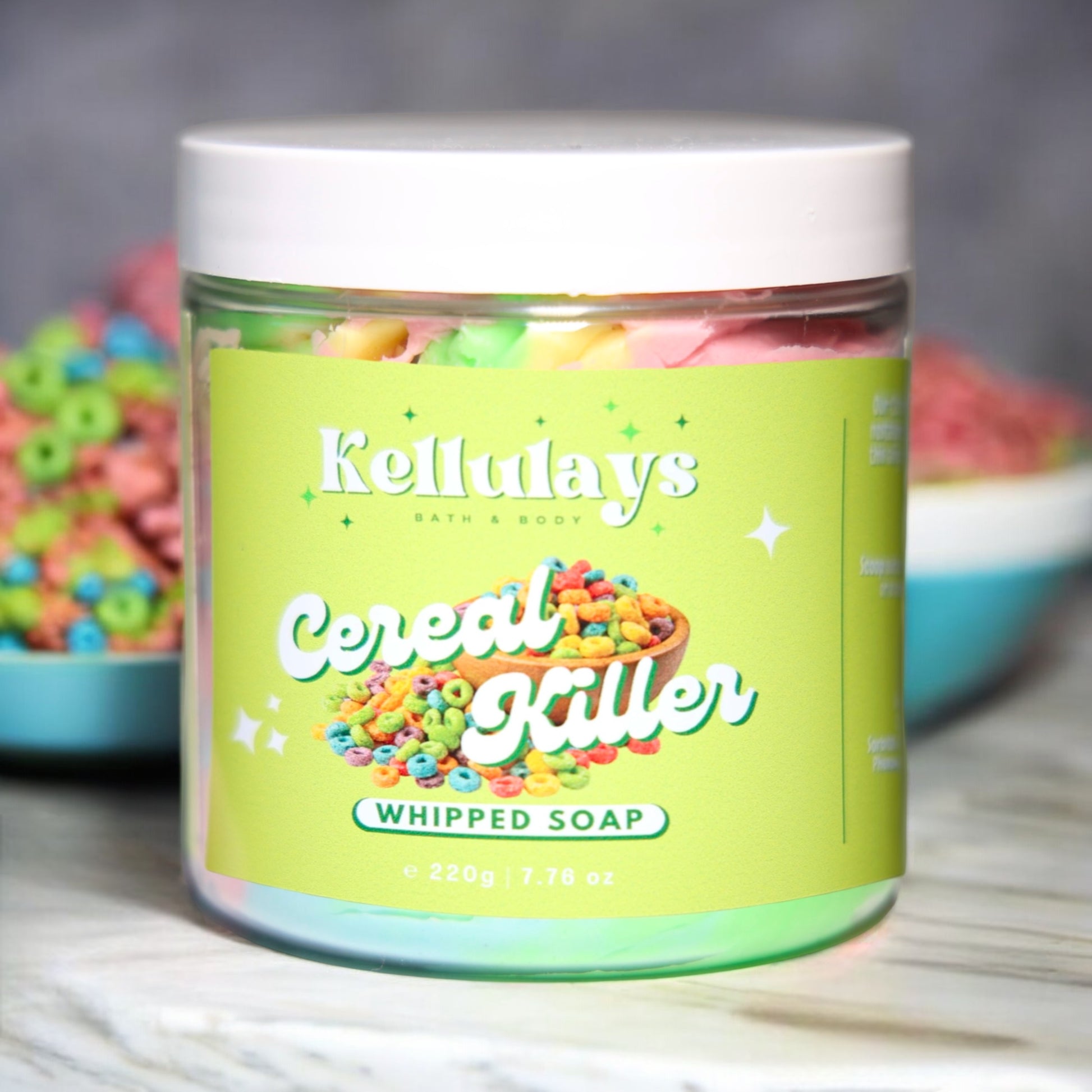 Image of Kellulays Cereal Killer Whipped Soap. The 220g jar features a bright green label with the product name 'Cereal Killer' in bold white letters and a colorful cereal graphic. Inside, the whipped soap is visible in vibrant pink, green, and yellow swirls. A bowl of colorful cereal loops is in the background, adding to the playful theme.