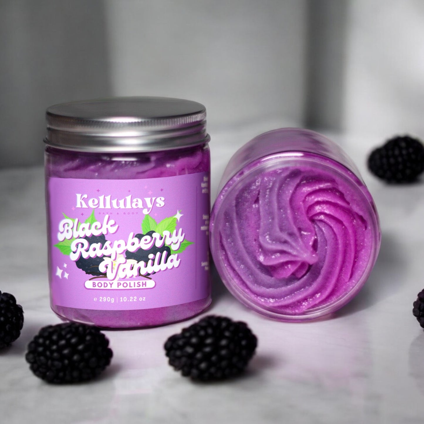 Two jars of Kellulays Black Raspberry Vanilla Body Polish, one upright and one open, showcasing its purple swirl texture. The packaging features a raspberry and vanilla design, with blackberries nearby.