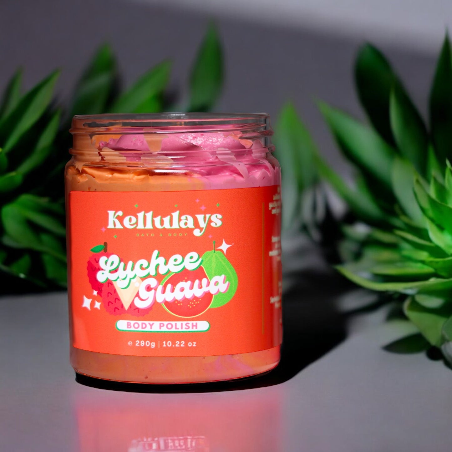 Jar of Kellulays Lychee Guava Body Polish with pink and orange swirls inside, set on a reflective surface with green succulents in the background, highlighting a vibrant tropical theme.