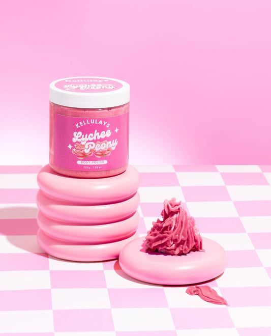 Jar of Kellulays Lychee Peony Body Polish on a pink and white checkered surface, with a pink whipped swirl next to it, creating a vibrant and stylish presentation.