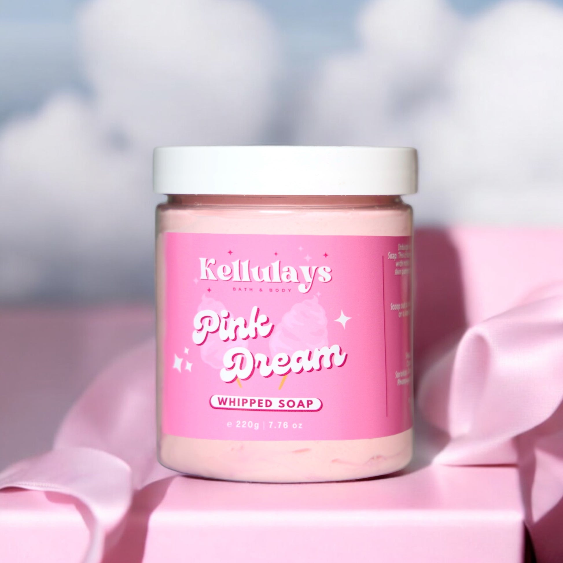 Image of Kellulays Bath & Body Pink Dream Whipped Soap in a clear jar with a white lid, sitting on a pink surface. The soap has a pink hue, and the label features a playful design with the product name and cotton candy graphics, evoking a sense of sweetness and indulgence.