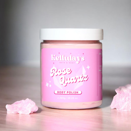 Image of Kellulays Rose Quartz Body Polish in a pink jar with a white lid. The label on the jar features the Kellulays logo and the product name in white, with subtle sparkles and a rose quartz crystal design in the background. The jar is placed on a wooden surface with pieces of pink rose quartz nearby, reflecting the product’s theme and aesthetic.
