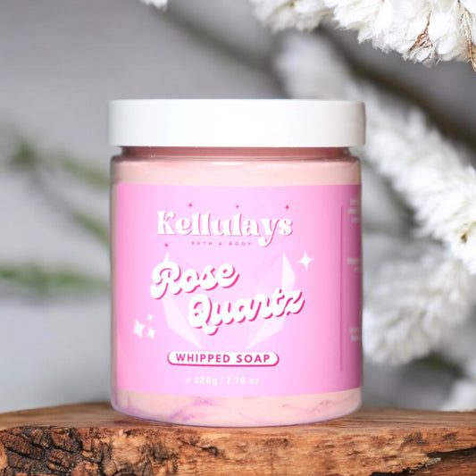 Kellulays Rose Quartz Whipped Soap. The 220g jar features a soft pink label with the product name ‘Rose Quartz’ in elegant white lettering and a subtle rose quartz graphic. The whipped soap inside is a delicate pink color. The jar is placed on a wooden surface, with a blurred background of fluffy white flowers, creating a serene and elegant setting.