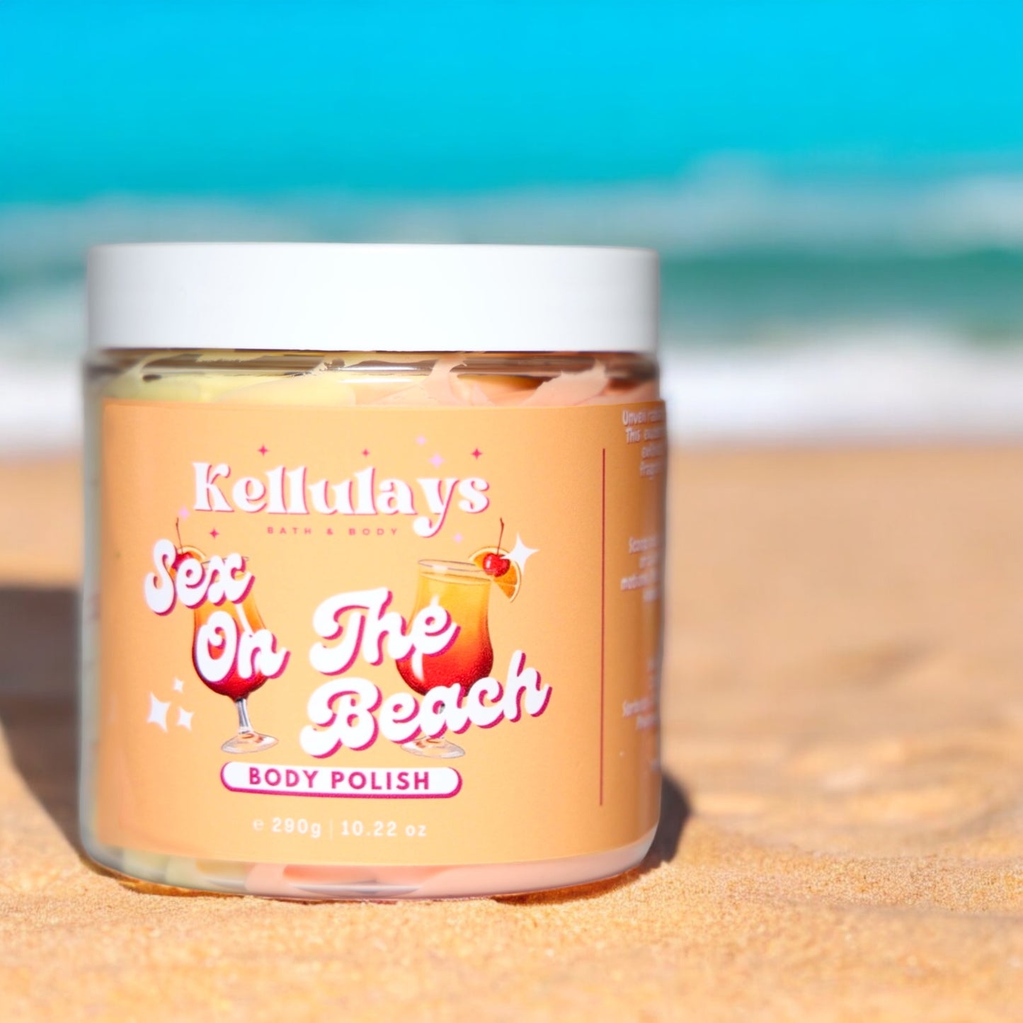 Jar of Kellulays ‘Sex On The Beach’ body polish on a sandy beach with ocean background, featuring a peach label with tropical cocktail illustrations.