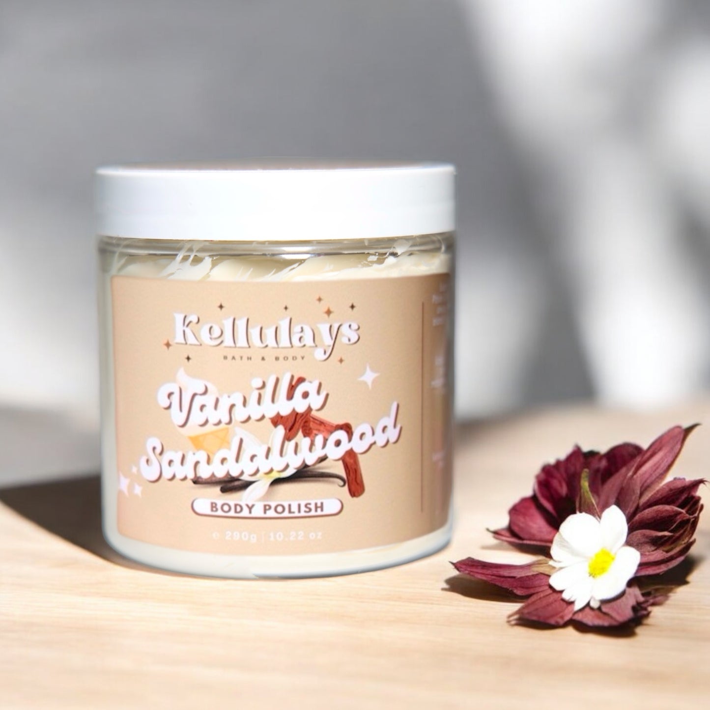 Jar of Kellulays Vanilla Sandalwood Body Polish on a wooden surface with decorative flowers, featuring a beige label and creamy texture inside, reflecting a soothing and luxurious product.