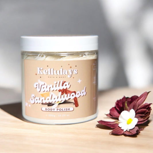 Jar of Kellulays Vanilla Sandalwood Body Polish on a wooden surface with decorative flowers, featuring a beige label and creamy texture inside, reflecting a soothing and luxurious product.