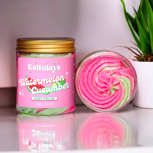 A jar of Kellulays Watermelon Cucumber Body Polish with a pink and green swirled texture visible inside, sitting on a white surface next to a small potted plant. The label features a vibrant pink background with the brand name and product details in playful, colorful fonts, showcasing the product’s fun and refreshing aesthetic.