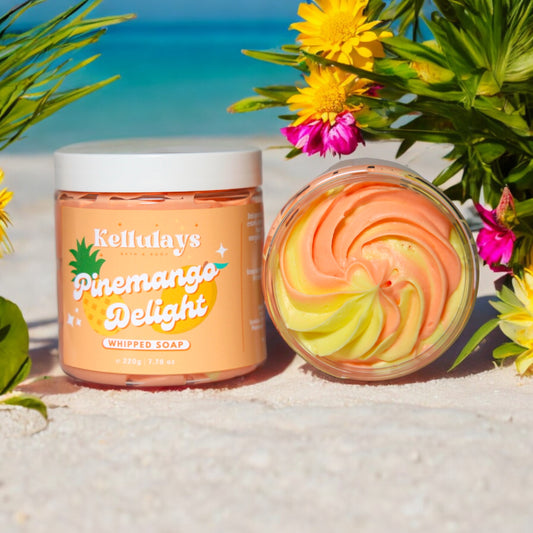 Kellulays Pinemango Delight Whipped Soap. The 220g jar is displayed on a sandy beach, featuring a tropical-themed label with the product name in white and orange lettering and a pineapple graphic. The whipped soap inside shows vibrant orange and yellow swirls. Surrounding the product are colourful tropical flowers and lush greenery, with the ocean visible in the background, creating a beachy, refreshing atmosphere.