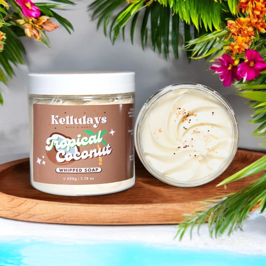 Kellulays Tropical Coconut Whipped Soap. The 220g jar has a brown label with the product name in white and green letters, decorated with a coconut graphic. The whipped soap inside is creamy white with a swirl pattern and a sprinkle of coconut flakes. The jar and open soap container are set on a wooden tray surrounded by tropical foliage and vibrant flowers, evoking a lush, island atmosphere.