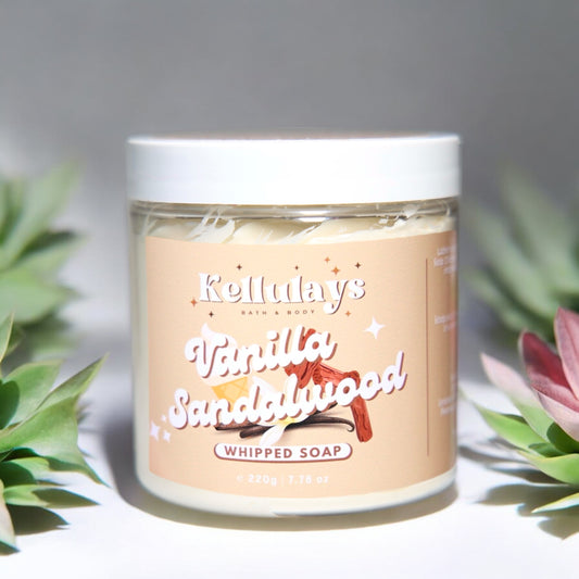 Image of a jar of Kellulays Bath & Body Vanilla Sandalwood Whipped Soap. The jar is labeled with a beige-colored sticker featuring the product name 'Vanilla Sandalwood' in bold, stylized white font with illustrations of vanilla pods and sandalwood pieces. The jar contains 220g (7.76 oz) of whipped soap, with a white lid. The background includes soft-focus green and pink succulents, adding a natural aesthetic to the presentation.