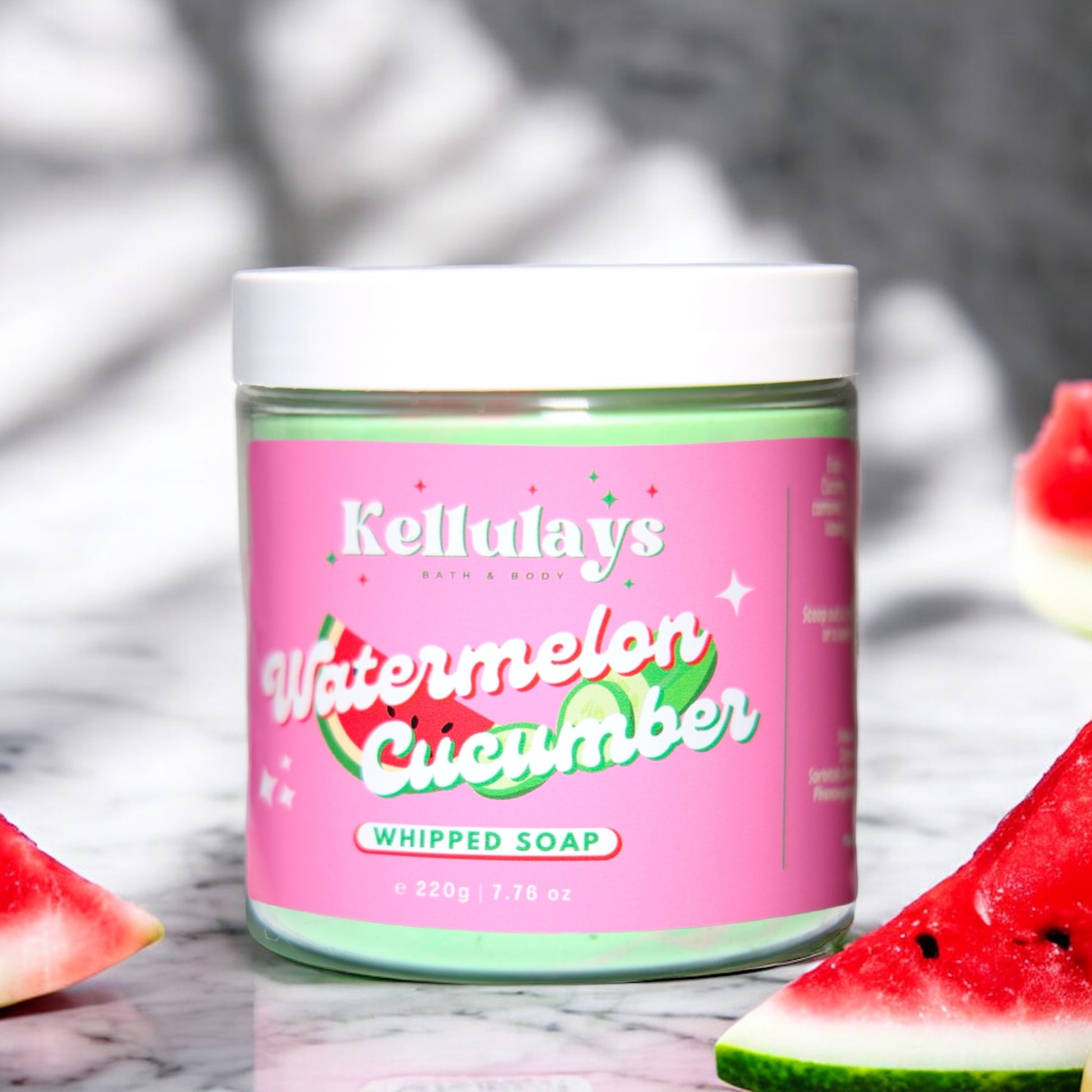 Kellulays Watermelon Cucumber Whipped Soap. The 220g jar has a vibrant pink label with the product name in white and green lettering, accented by graphics of watermelon slices and cucumber. The whipped soap inside is a refreshing green color. Slices of watermelon and cucumber are artfully placed around the jar, emphasizing the fresh and fruity theme. The background is a light marble surface, enhancing the bright and clean aesthetic.