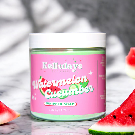 Kellulays Watermelon Cucumber Whipped Soap. The 220g jar has a vibrant pink label with the product name in white and green lettering, accented by graphics of watermelon slices and cucumber. The whipped soap inside is a refreshing green color. Slices of watermelon and cucumber are artfully placed around the jar, emphasizing the fresh and fruity theme. The background is a light marble surface, enhancing the bright and clean aesthetic.