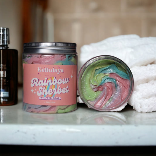 A jar of Kellulays Rainbow Sherbet Whipped Soap, showcasing a colourful rainbow swirl with vibrant hues of pink, yellow, blue, and green. The handmade bath whip is infused with nourishing cocoa and shea butters, offering a silky texture designed to cleanse, moisturize, and rejuvenate the skin. The label on the jar emphasizes the fun and fruity scent reminiscent of classic rainbow sherbet, perfect for adding a playful touch to your bathing routine.