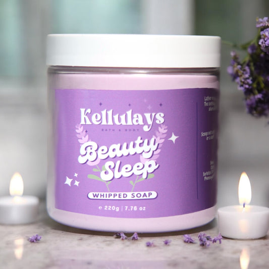 Image of a jar of Kellulays Bath & Body Beauty Sleep Whipped Soap. The jar is labeled with a purple sticker featuring the product name 'Beauty Sleep' in bold white font, accompanied by lavender and botanical illustrations. The jar contains 220g (7.76 oz) of whipped soap and has a white lid. The background includes soft-focus lavender sprigs and lit candles, creating a calming and serene atmosphere.