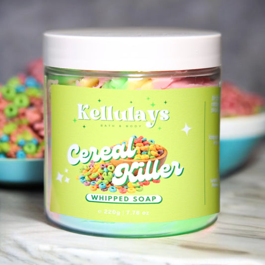 Image of Kellulays Cereal Killer Whipped Soap. The 220g jar features a bright green label with the product name 'Cereal Killer' in bold white letters and a colorful cereal graphic. Inside, the whipped soap is visible in vibrant pink, green, and yellow swirls. A bowl of colorful cereal loops is in the background, adding to the playful theme.