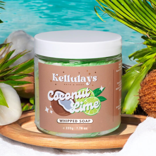 A jar of Kellulays Coconut Lime Whipped Soap, featuring a brown label with coconut and lime graphics. The jar is filled with a vibrant green, white, yellow soap and has a white lid. It is placed on a wooden tray surrounded by tropical elements, including a coconut, palm leaves, and a pool of blue water in the background, creating a refreshing and tropical setting.