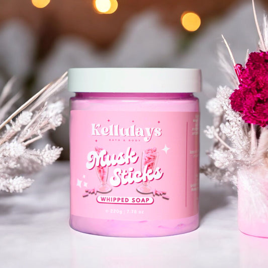 Image of a jar of Kellulays Musk Sticks Whipped Soap. The jar is pink with white lettering, featuring an illustration of musk stick candies. It contains 220g (7.76 oz) of product. The jar is placed on a soft pink surface, with decorative white and magenta flowers surrounding it. Soft, warm lights are visible in the blurred background, creating a cozy ambiance.