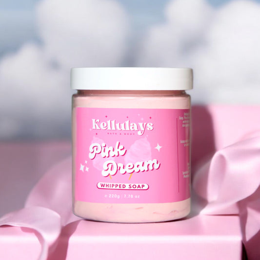 Image of Kellulays Bath & Body Pink Dream Whipped Soap in a clear jar with a white lid, sitting on a pink surface. The soap has a pink hue, and the label features a playful design with the product name and cotton candy graphics, evoking a sense of sweetness and indulgence.