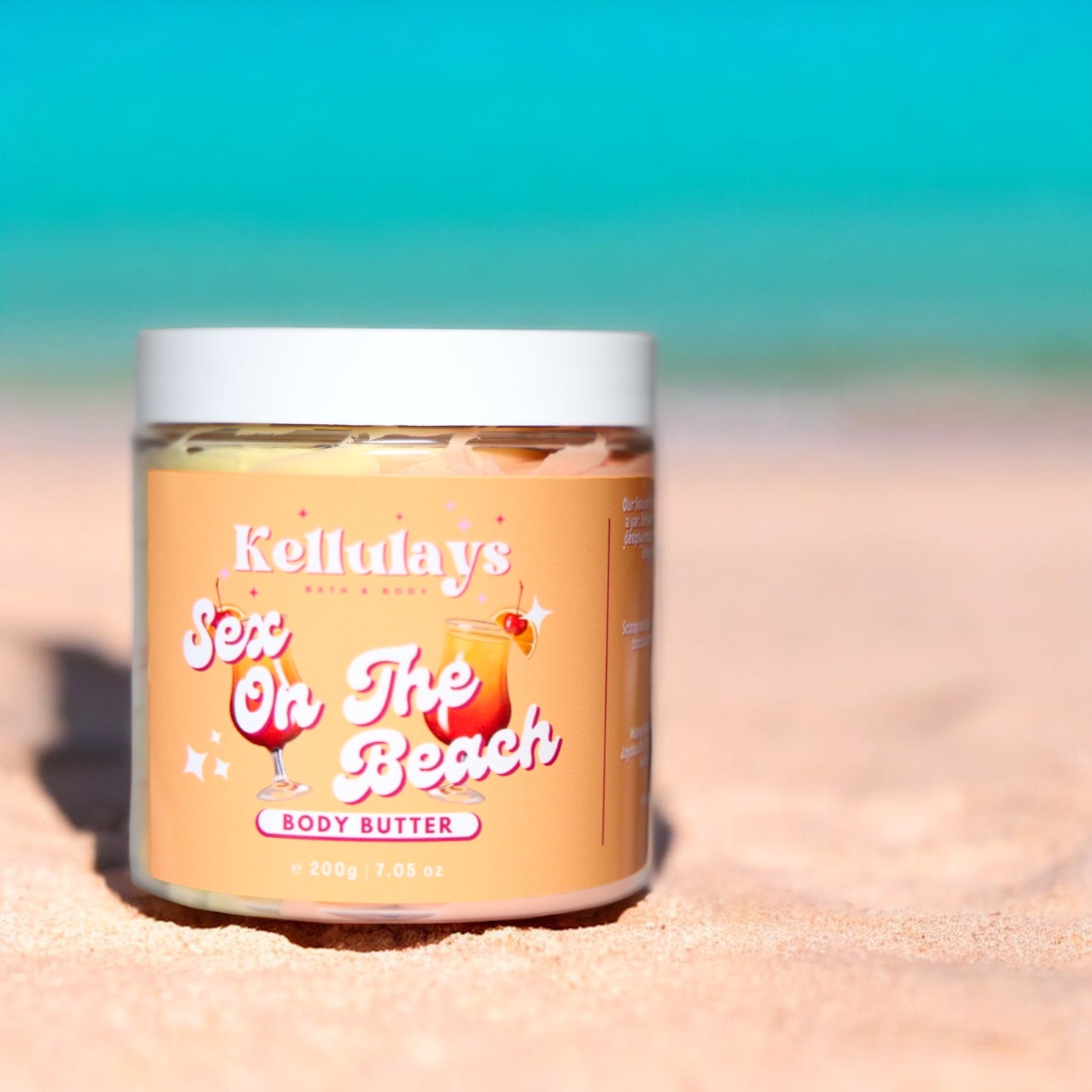 Close-up of a Kellulays Bath & Body ‘Sex On The Beach’ body butter jar resting on a sandy beach. The jar is labeled with vibrant, playful typography and features illustrations of tropical cocktails. The turquoise ocean and blue sky provide a serene backdrop, enhancing the summery, beach-themed aesthetic of the product.