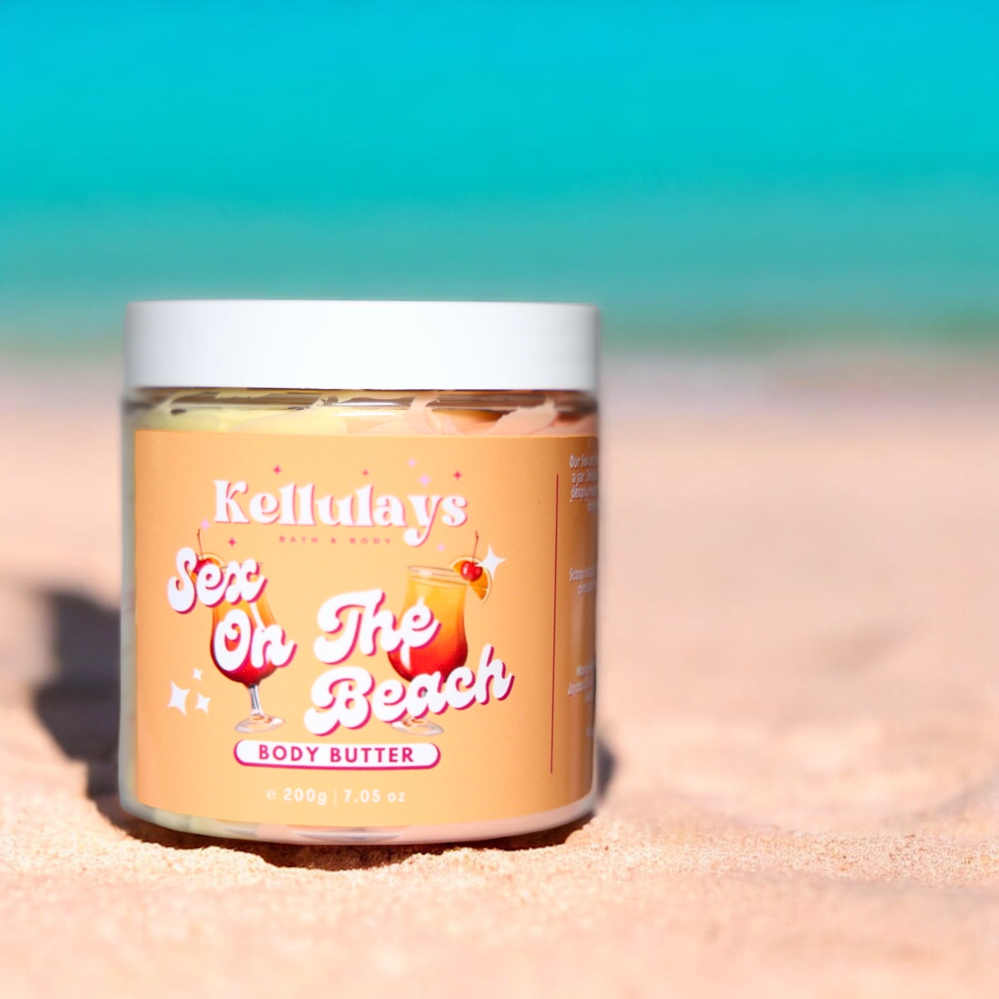 Close-up of a Kellulays Bath & Body ‘Sex On The Beach’ body butter jar resting on a sandy beach. The jar is labeled with vibrant, playful typography and features illustrations of tropical cocktails. The turquoise ocean and blue sky provide a serene backdrop, enhancing the summery, beach-themed aesthetic of the product.