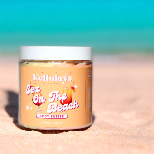 Close-up of a Kellulays Bath & Body ‘Sex On The Beach’ body butter jar resting on a sandy beach. The jar is labeled with vibrant, playful typography and features illustrations of tropical cocktails. The turquoise ocean and blue sky provide a serene backdrop, enhancing the summery, beach-themed aesthetic of the product.