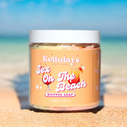 Image of Kellulays Bath & Body Sex on the Beach Whipped Soap in a clear jar with a white lid, placed on a sandy beach with a blurred ocean backdrop. The label features a playful cocktail graphic, highlighting the tropical and indulgent nature of this handmade bath whip, enriched with shea and cocoa butters.