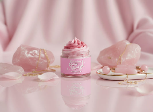 A luxurious jar of Kellulays Rose Quartz Body Butter sits surrounded by rose quartz crystals, soft pink petals, and gold jewellery on a glossy pastel pink surface. The whipped pink swirl inside the jar reflects a romantic, crystal inspired aesthetic, showcasing the product's creamy texture ad self-care appeal.