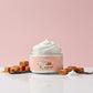 A jar of Kellulays Vanilla Caramel whipped body butter sitting on a glossy white surface, surrounded by soft caramel cubes and vanilla beans, with a pastel pink background. The jar is open, showing a smooth white whipped swirl, giving a dessert inspired luxurious skincare aesthetic.