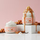 Two jars of Kellulays Vanilla Caramel whipped body butter styled against a soft pink background. One jar features a smooth white swirl, while the other is topped with a tall whipped peak covered in dripping caramel sauce. Caramel cubes and vanilla beans surround the jars for dessert inspired skincare aesthetic