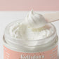 A close up of Kellulays Vanilla Caramel whipped body butter, showing fluffy, cloud like swirl being scooped with a spatula against a soft pink background. The creamy texture highlights the products light, whipped consistency.