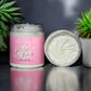A jar of Kellulays Plain Jane Body Butter with a pink label, featuring the product name in white script. The body butter has a creamy, smooth texture and is unscented. The open jar is displayed alongside a closed jar, surrounded by small green succulent plants, against a dark, reflective surface.